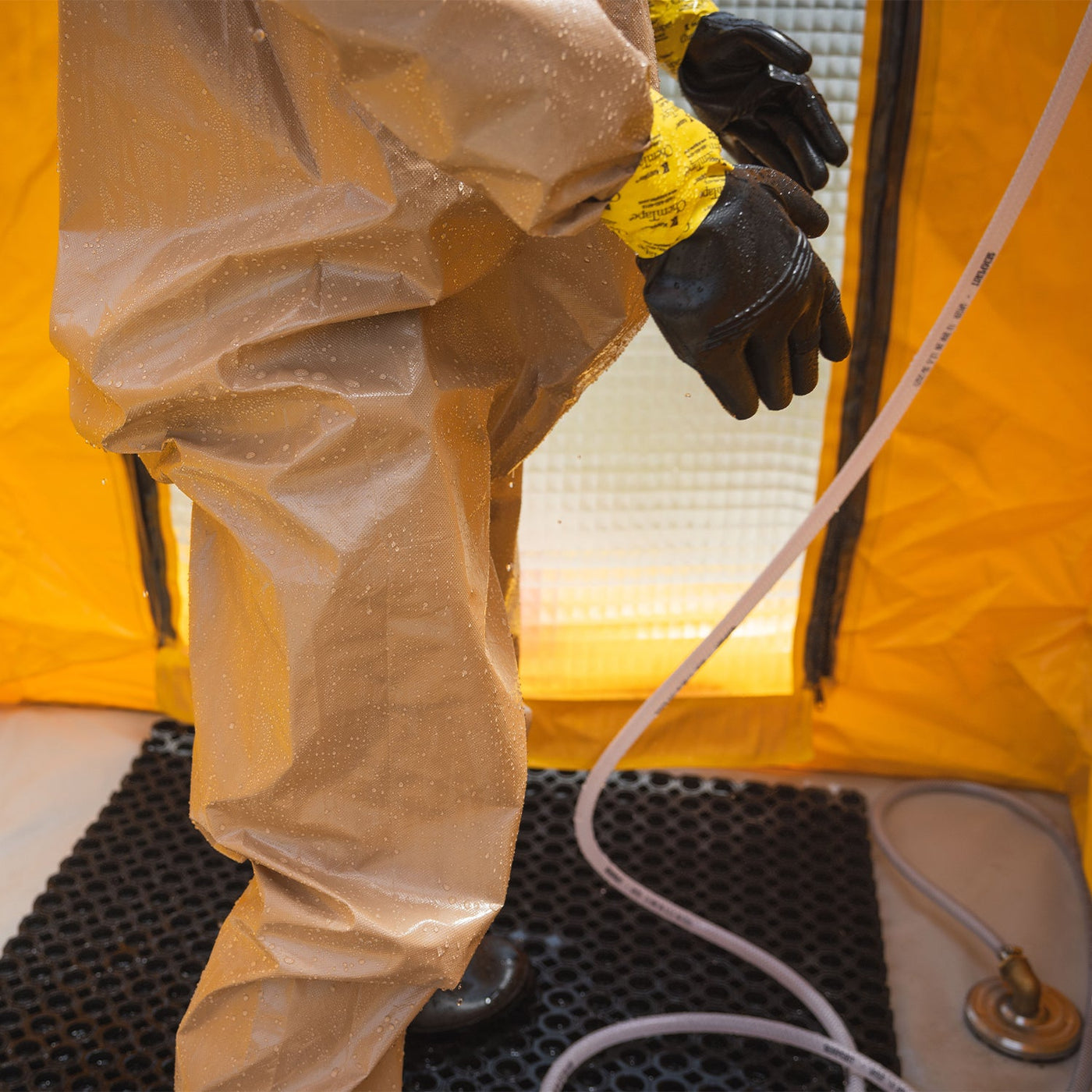 View of a man wearing a hazmat suit standing inside of the decontamination shower. The shower's floor can be seen, along with the drainage mat and a water disposal outlet.