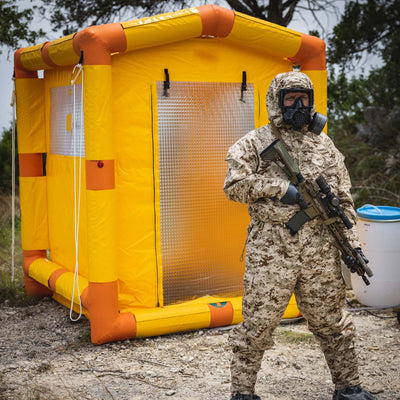 A soldier wearing a MOPP suit is guarding the decontamination shower with an AR-15 rifle.